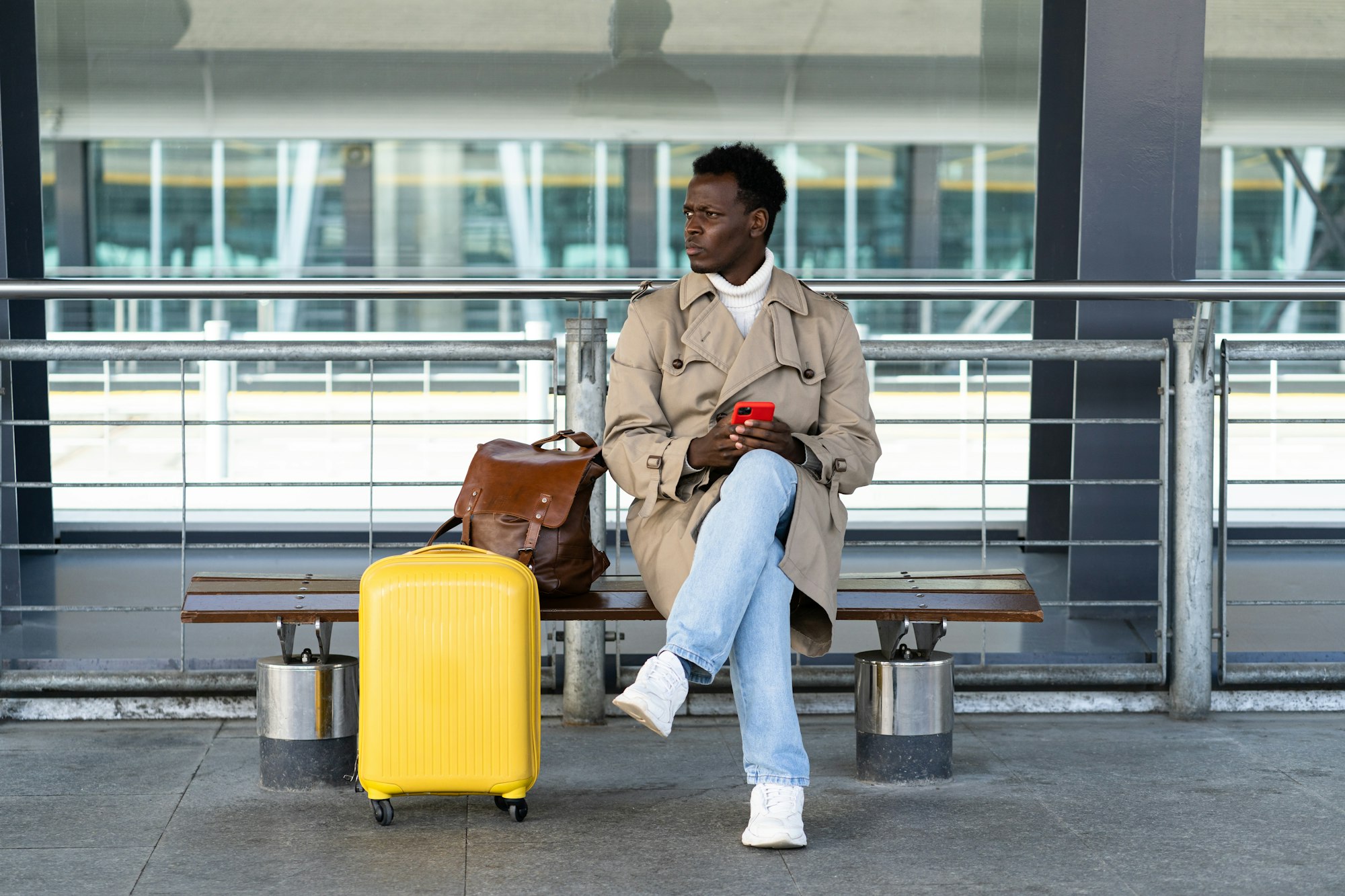 Black traveler man with suitcase sitting on bench in airport terminal, calling and looking for buffalo airport taxi